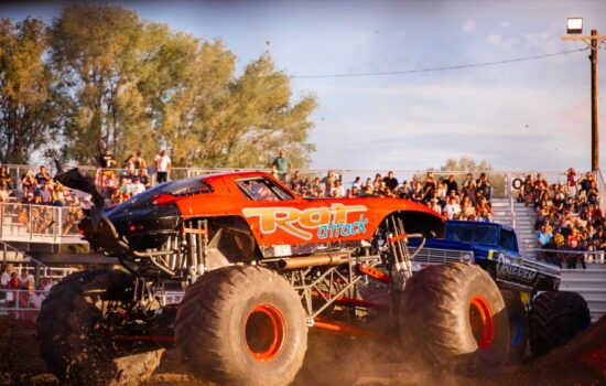 Two monster vehicles drive through mud with bleachers and fans in the background.